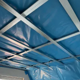 Stelaż z profili stalowych podwieszony do sufitu, zabezpieczony niebieską folią budowlaną, widoczny fragment okna z białymi ramami.