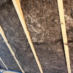 Izolacja z wełny mineralnej zamontowana między krokwiami poddasza, widoczne drewniane elementy konstrukcyjne i fragment instalacji elektrycznej.