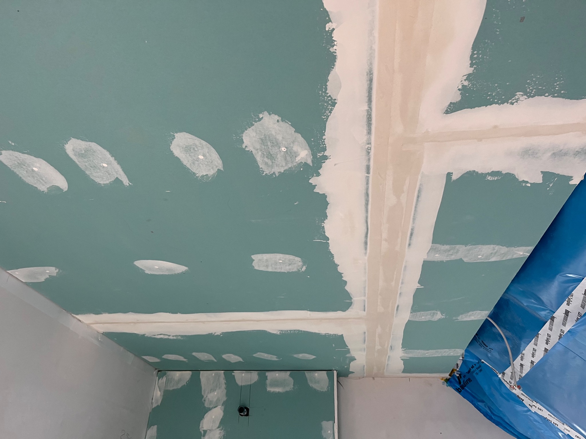 Niebieska folia zabezpieczająca w rogu pokoju, widok na sufit z płyt kartonowo-gipsowych, wstępnie wyszpachlowane łączenia i ślady po wkrętach.