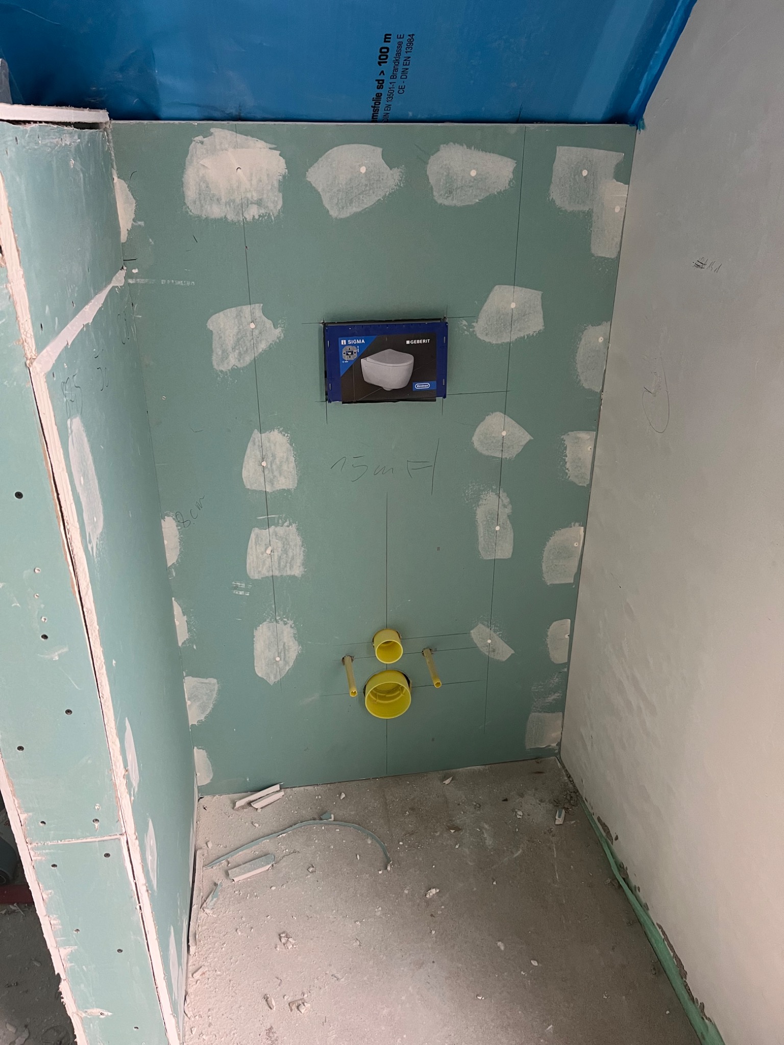 Montaż stelaża podtynkowego w łazience, widoczne płyty gipsowo-kartonowe z naniesionymi liniami pomocniczymi i punktami szpachlowania, żółte elementy instalacji wodnej i niebieski karton stelaża.