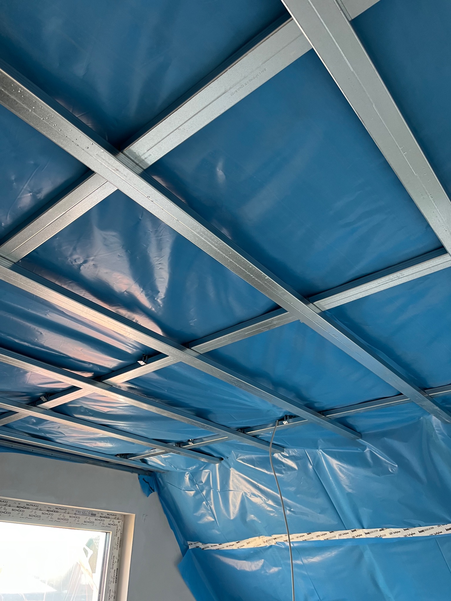 Stelaż z profili stalowych podwieszony do sufitu, zabezpieczony niebieską folią budowlaną, widoczny fragment okna z białymi ramami.