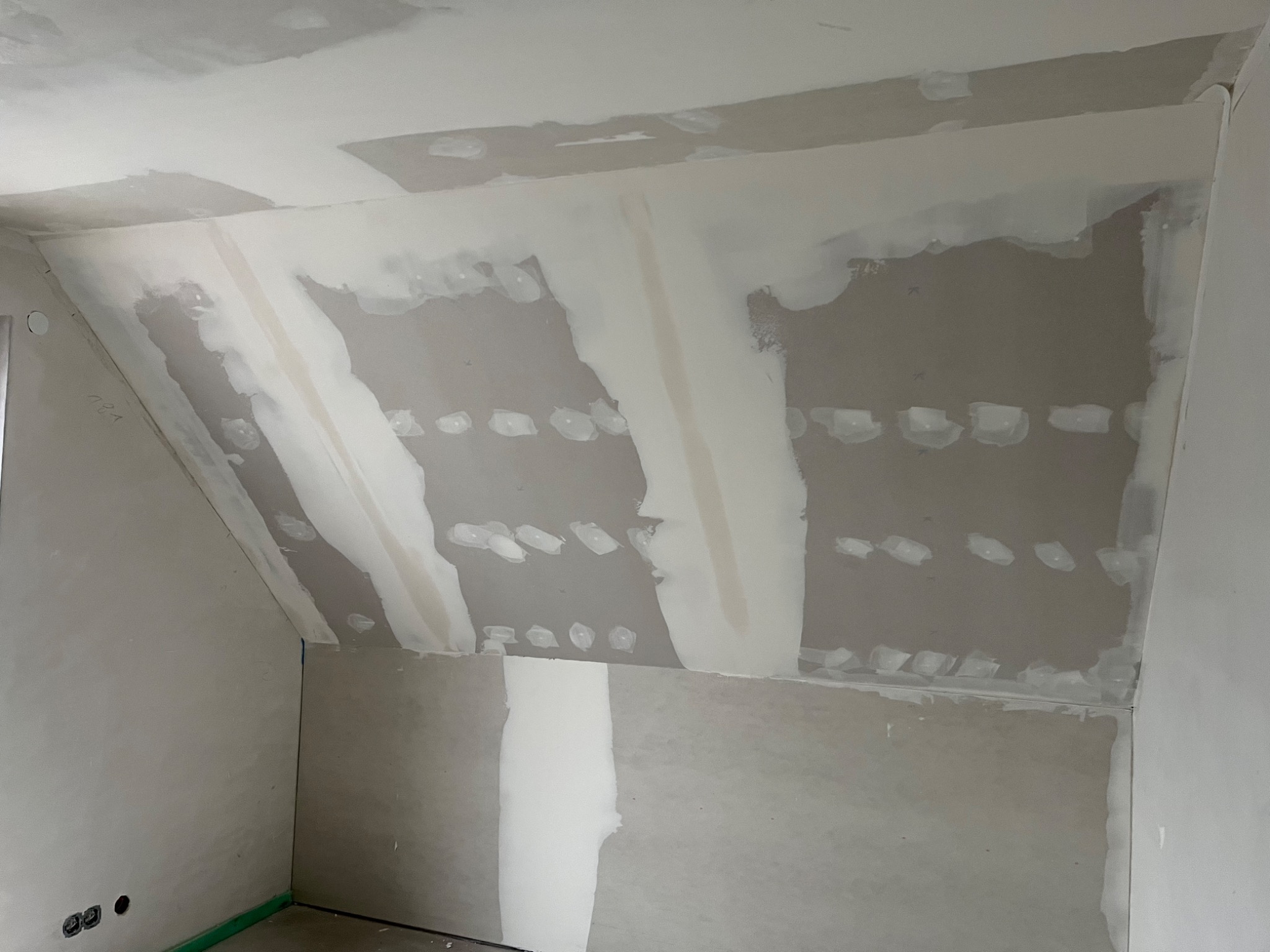 Poddasze ze skośnym sufitem w trakcie szpachlowania płyt karton-gips, widoczne łączenia wzmocnione taśmą i pokryte masą szpachlową, ściany częściowo przygotowane do dalszych prac wykończeniowych.