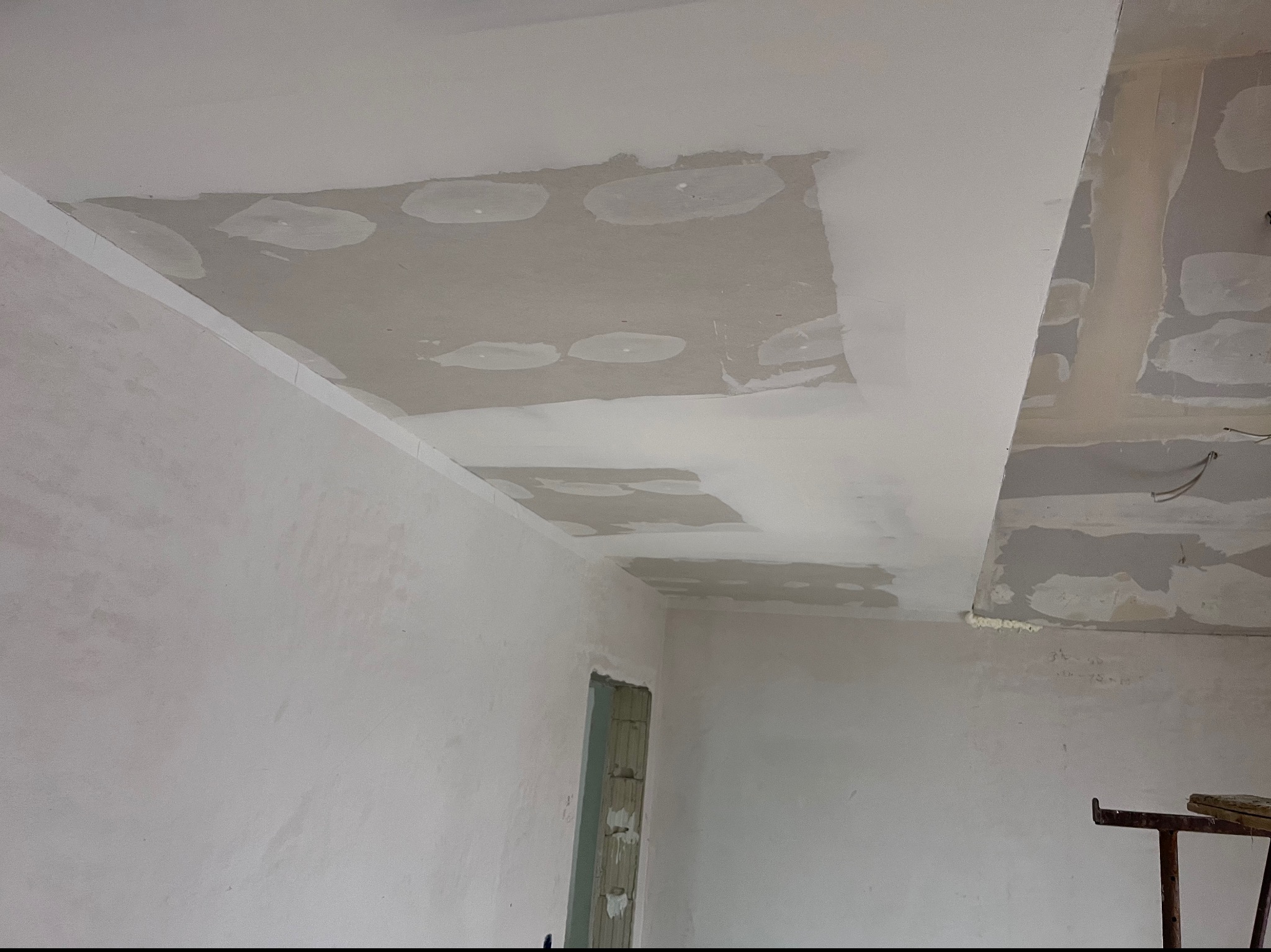 Wnętrze w trakcie remontu: fragment sufitu z widocznymi łatami szpachlowymi i świeżo pomalowaną powierzchnią, widoczne ślady po wcześniejszych pracach oraz fragment ściany z ościeżnicą.