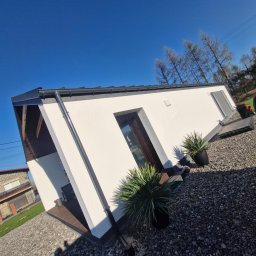 Art Prestige House - Nowoczesny, parterowy dom z białymi ścianami i ciemną stolarką okienną, otoczony szarym żwirem i roślinami w donicach, widziany z nietypowej perspektywy.