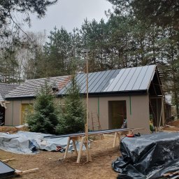 Art Prestige House - Budowa domu w lesie: częściowo pokryty blachą dach, otynkowana elewacja, widoczne rusztowania i narzędzia, drzewa iglaste w tle.