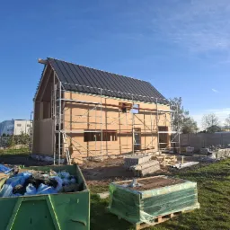 Wznoszony dom szkieletowy z czarnym dachem, na tle błękitnego nieba. Widoczne rusztowania i kontener na odpady budowlane. Faza budowy z elementami wykończeniowymi.