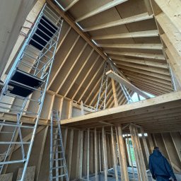 Art Prestige House - Wnętrze domu w trakcie budowy szkieletowej: widoczne drewniane elementy konstrukcyjne, rusztowania, surowe ściany z płyt OSB, wysoki strop z oknami na poddaszu.