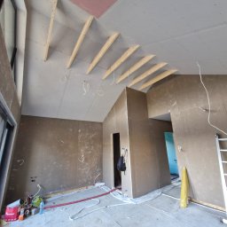 Art Prestige House - Wnętrze w trakcie wykończenia: surowe ściany z widocznymi instalacjami, drewniana konstrukcja dachu, okno i drabina. Etap prac wykończeniowych przed malowaniem.