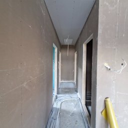Art Prestige House - Korytarz w trakcie wykańczania: surowe ściany z płyt gipsowych, widoczne kable elektryczne na podłodze, przygotowane otwory na drzwi. W tle widoczny fragment niebieskiej ściany.
