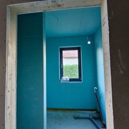 Art Prestige House - Wnętrze w trakcie prac wykończeniowych: ściany obłożone płytami gipsowo-kartonowymi, widoczne okno z widokiem na zieleń, instalacja sanitarna na podłodze.