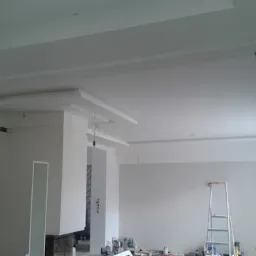 Wykończony sufit podwieszany z dekoracyjnymi elementami w jasnym, przestronnym wnętrzu w trakcie remontu, widoczna drabina i narzędzia.