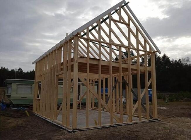 Konstrukcja szkieletowa domu w trakcie budowy, widoczne drewniane elementy nośne, szkielet dachu i ścian. W tle zalesiony teren i przyczepa kempingowa.