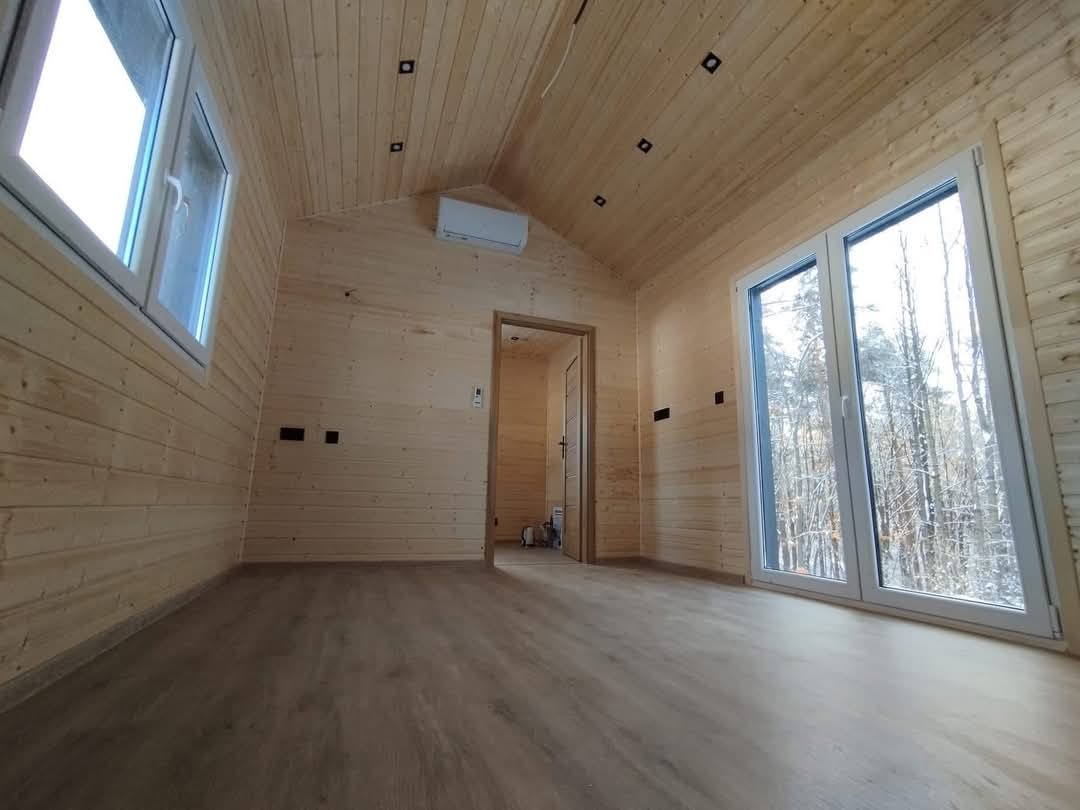 Jasne, drewniane wnętrze domu z oknami na las. Widoczne panele podłogowe, drewniane ściany i sufit ze światłami. Drzwi w głębi prowadzą do kolejnego pomieszczenia.