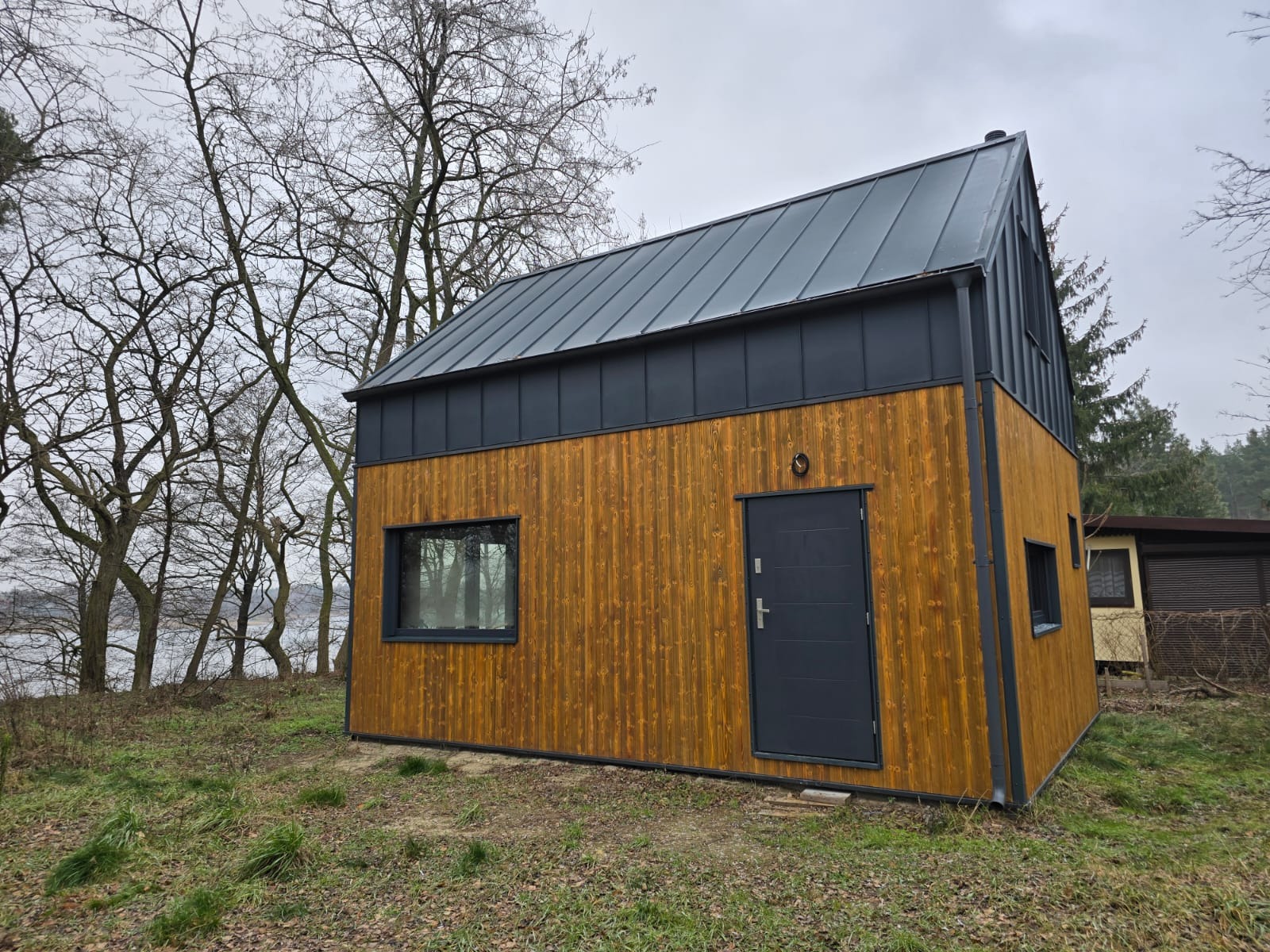 Nowoczesny dom szkieletowy z drewnianą elewacją i ciemnoszarą blachą na dachu, otoczony drzewami i trawą. Widok na jezioro w tle.