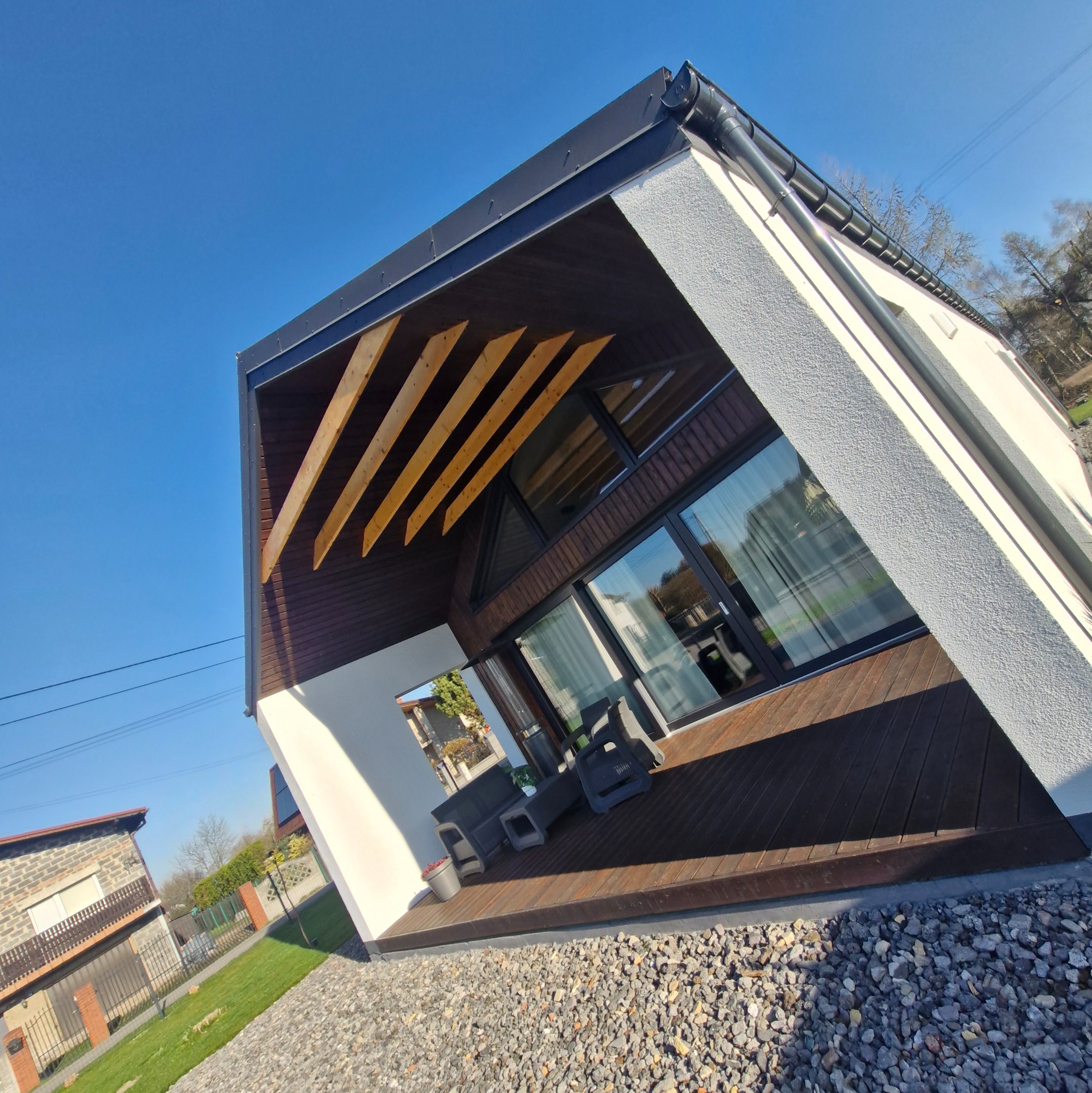 Nowoczesny dom z drewnianym tarasem i jasną elewacją, widziany z perspektywy żabiej. Na pierwszym planie szara nawierzchnia z kamienia.