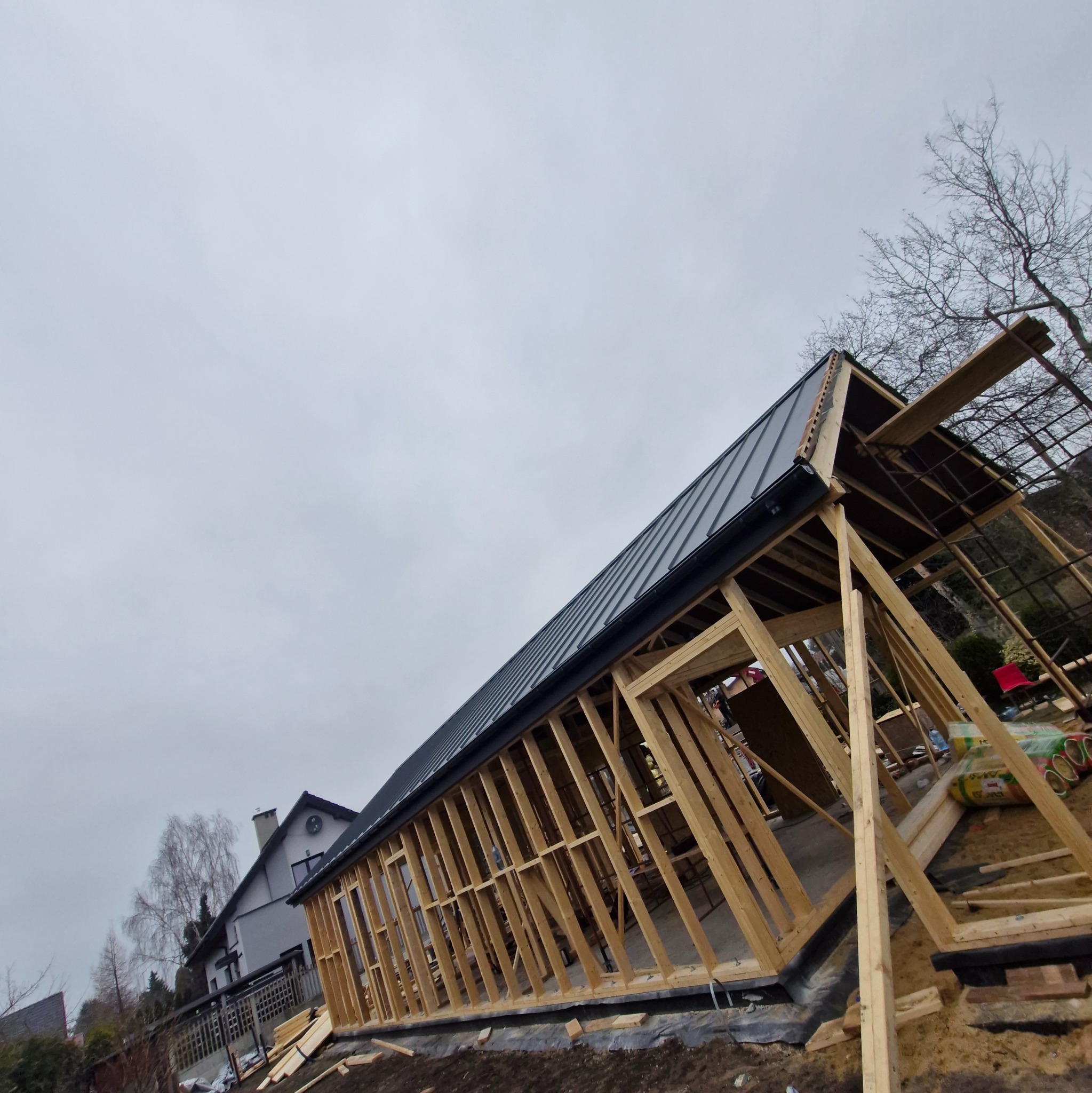 Szkielet domu w budowie z widocznym zadaszeniem i elementami konstrukcyjnymi. Ujęcie z dołu, pochmurne niebo w tle.
