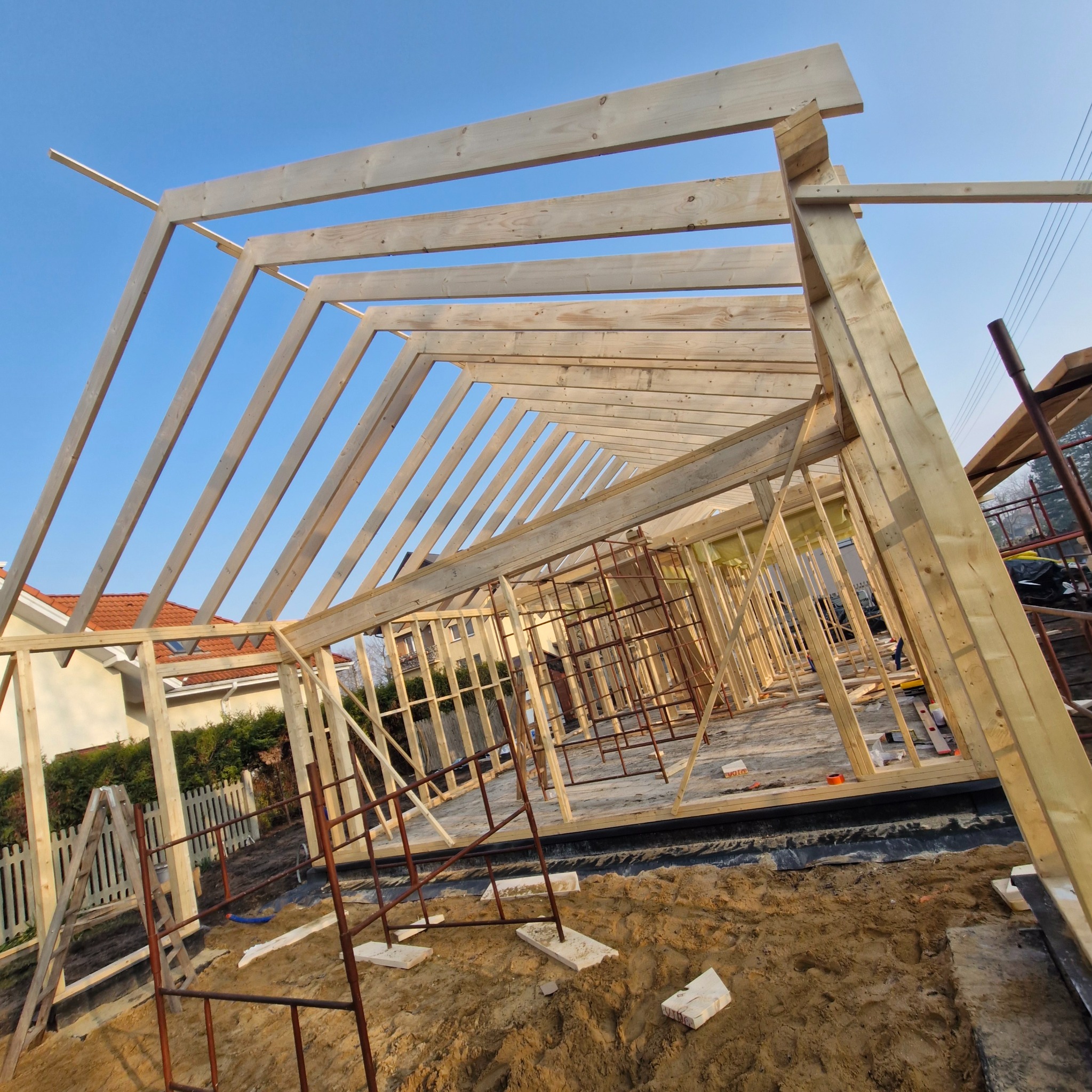 Drewniany szkielet domu w trakcie budowy, widziany z dołu pod kątem, z elementami rusztowania i sąsiednimi domami w tle, na tle błękitnego nieba.