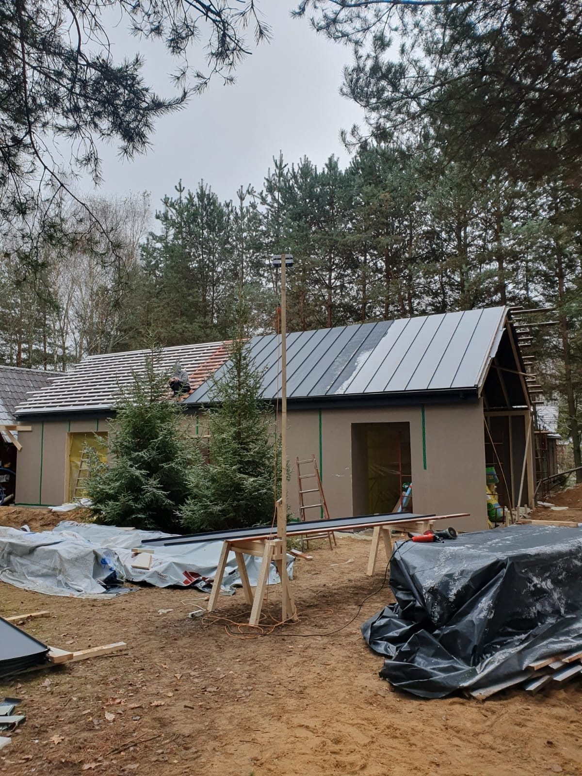 Budowa domu w lesie: częściowo pokryty blachą dach, otynkowana elewacja, widoczne rusztowania i narzędzia, drzewa iglaste w tle.
