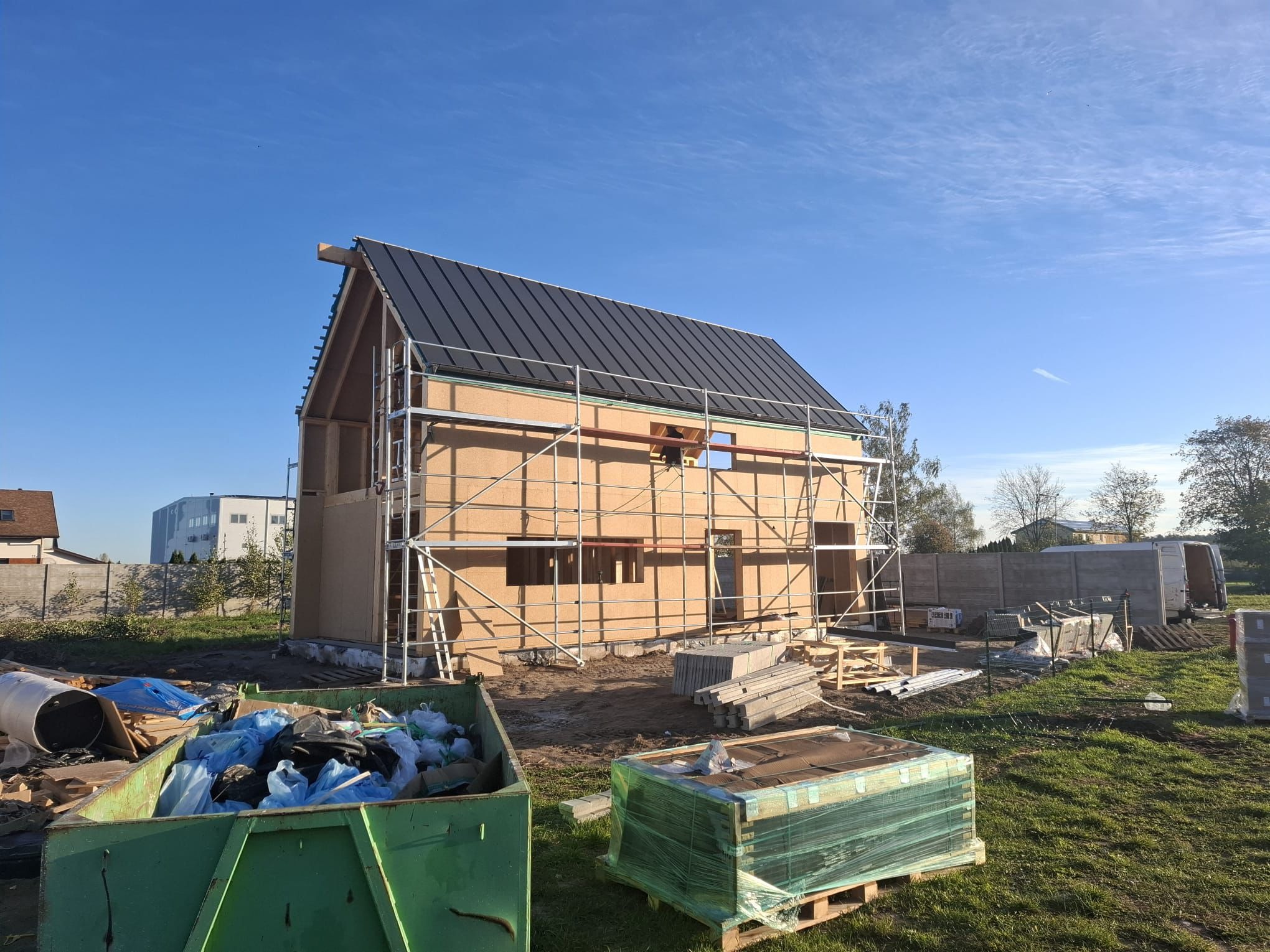 Wznoszony dom szkieletowy z czarnym dachem, na tle błękitnego nieba. Widoczne rusztowania i kontener na odpady budowlane. Faza budowy z elementami wykończeniowymi.
