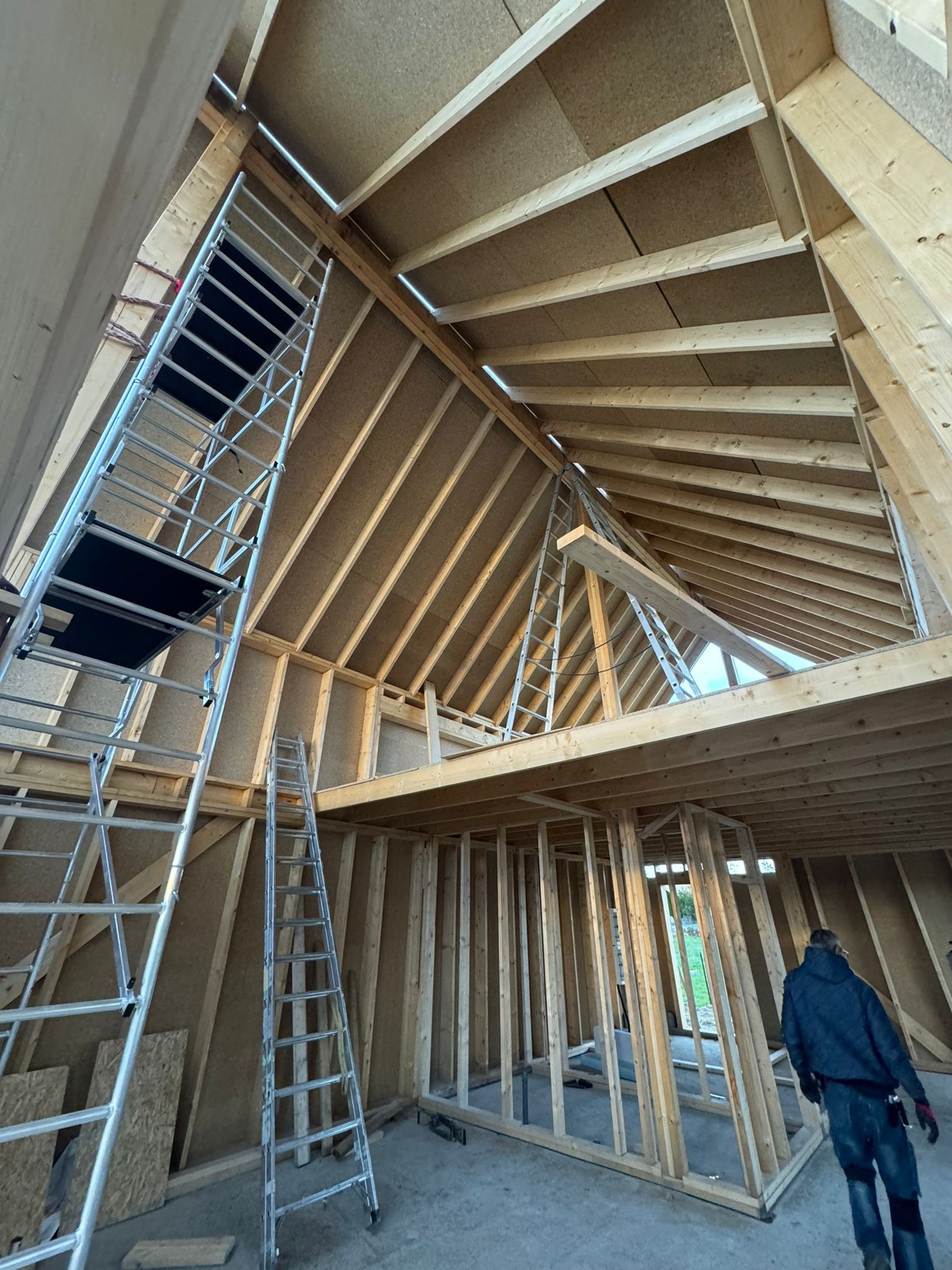 Wnętrze domu w trakcie budowy szkieletowej: widoczne drewniane elementy konstrukcyjne, rusztowania, surowe ściany z płyt OSB, wysoki strop z oknami na poddaszu.