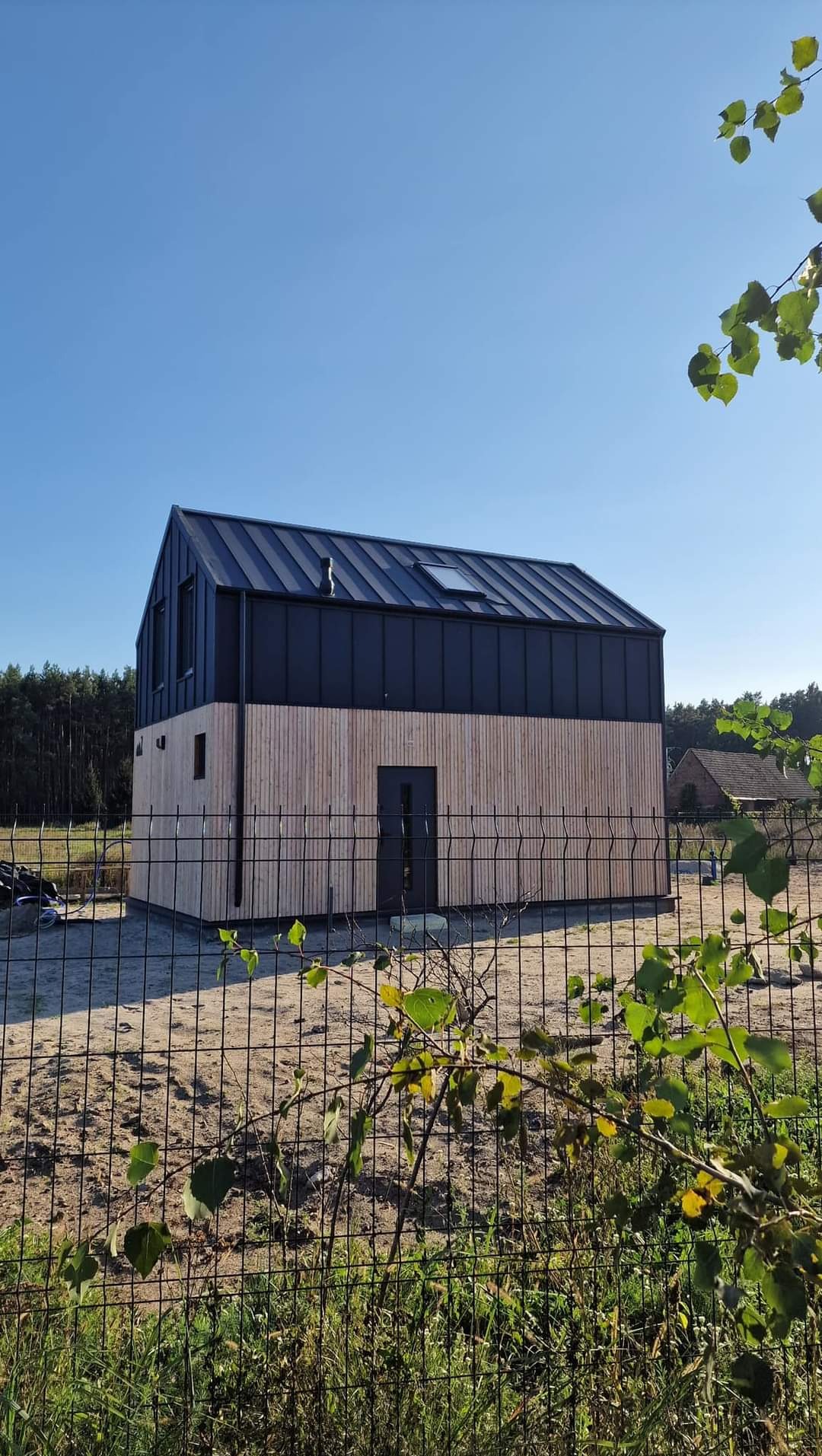 Nowoczesny dom szkieletowy z drewnianą elewacją i ciemnym dachem, widoczny przez ogrodzenie z siatki. Błękitne niebo w tle.