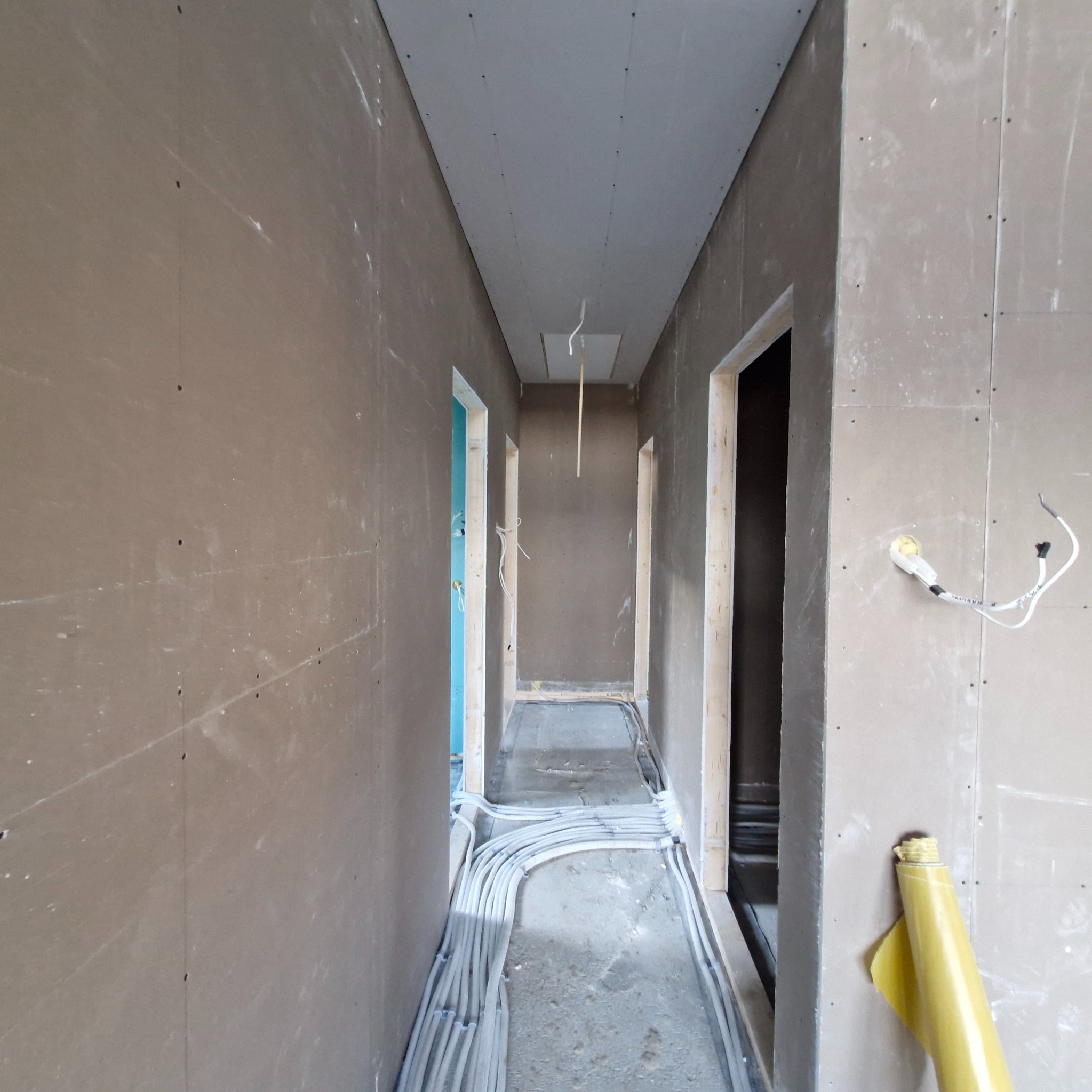 Korytarz w trakcie wykańczania: surowe ściany z płyt gipsowych, widoczne kable elektryczne na podłodze, przygotowane otwory na drzwi. W tle widoczny fragment niebieskiej ściany.