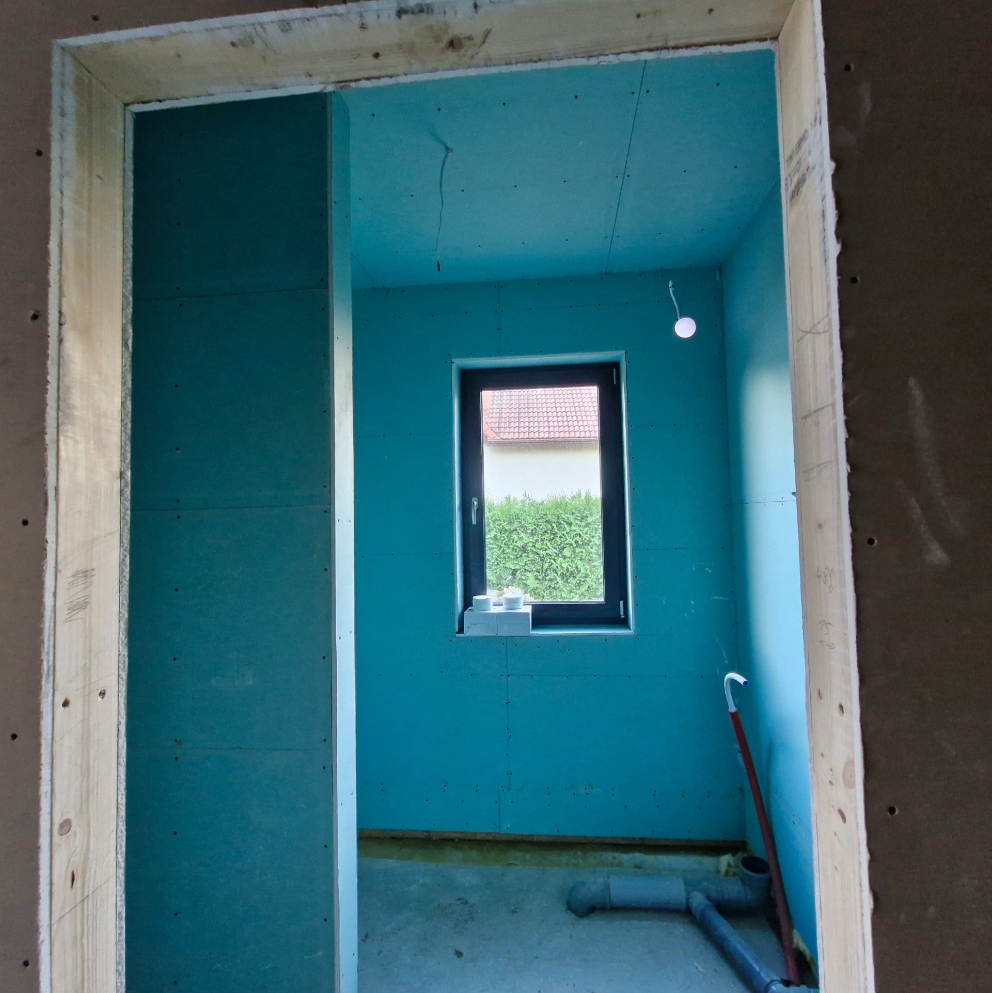 Wnętrze w trakcie prac wykończeniowych: ściany obłożone płytami gipsowo-kartonowymi, widoczne okno z widokiem na zieleń, instalacja sanitarna na podłodze.