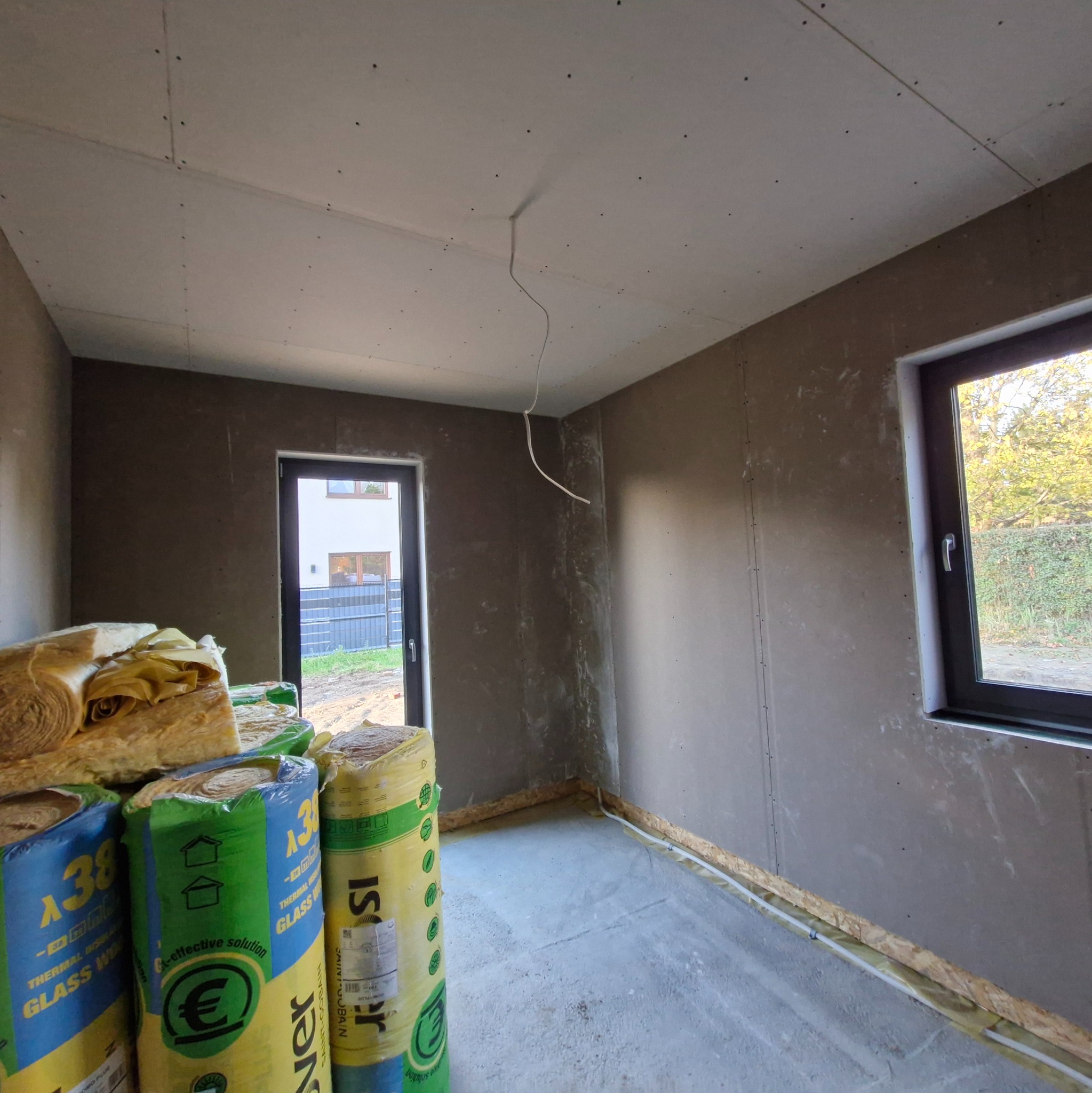 Wnętrze w trakcie prac wykończeniowych: ściany przygotowane do szpachlowania, widoczna izolacja termiczna w rolkach i wystający przewód elektryczny.