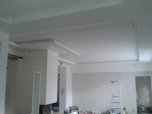 Wykończony sufit podwieszany z dekoracyjnymi elementami w jasnym, przestronnym wnętrzu w trakcie remontu, widoczna drabina i narzędzia.