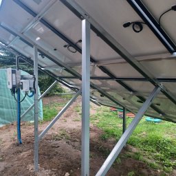 VOLT-MAR usługi elektryczne Marek Kierepka - Widok od spodu na panele słoneczne zamontowane na metalowej konstrukcji, z widocznym inwerterem i okablowaniem, na tle ogrodu z tunelem foliowym.