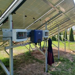 VOLT-MAR usługi elektryczne Marek Kierepka - Widok od dołu na panele fotowoltaiczne zamontowane na metalowej konstrukcji, z widocznymi puszkami przyłączeniowymi i inwerterem Hypontech, w tle trawnik i tuje, na jednym ze słupków konstrukcji...