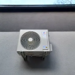 Biała jednostka zewnętrzna klimatyzatora marki Gree zamontowana na jasnoszarej elewacji dwukondygnacyjnego budynku z czarną rynną.