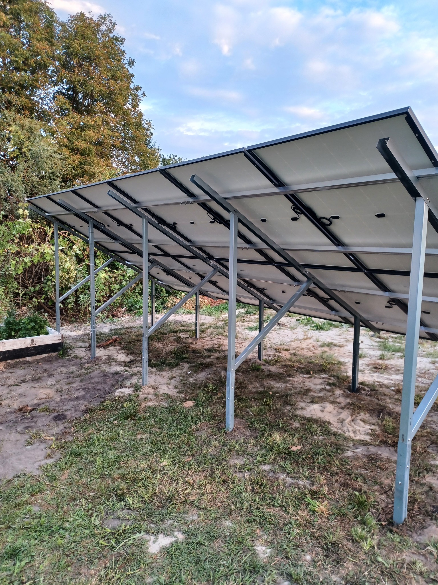 Widok od spodu na panele słoneczne zamontowane na metalowej konstrukcji, widoczne przewody i elementy mocujące, w tle drzewa i pochmurne niebo.