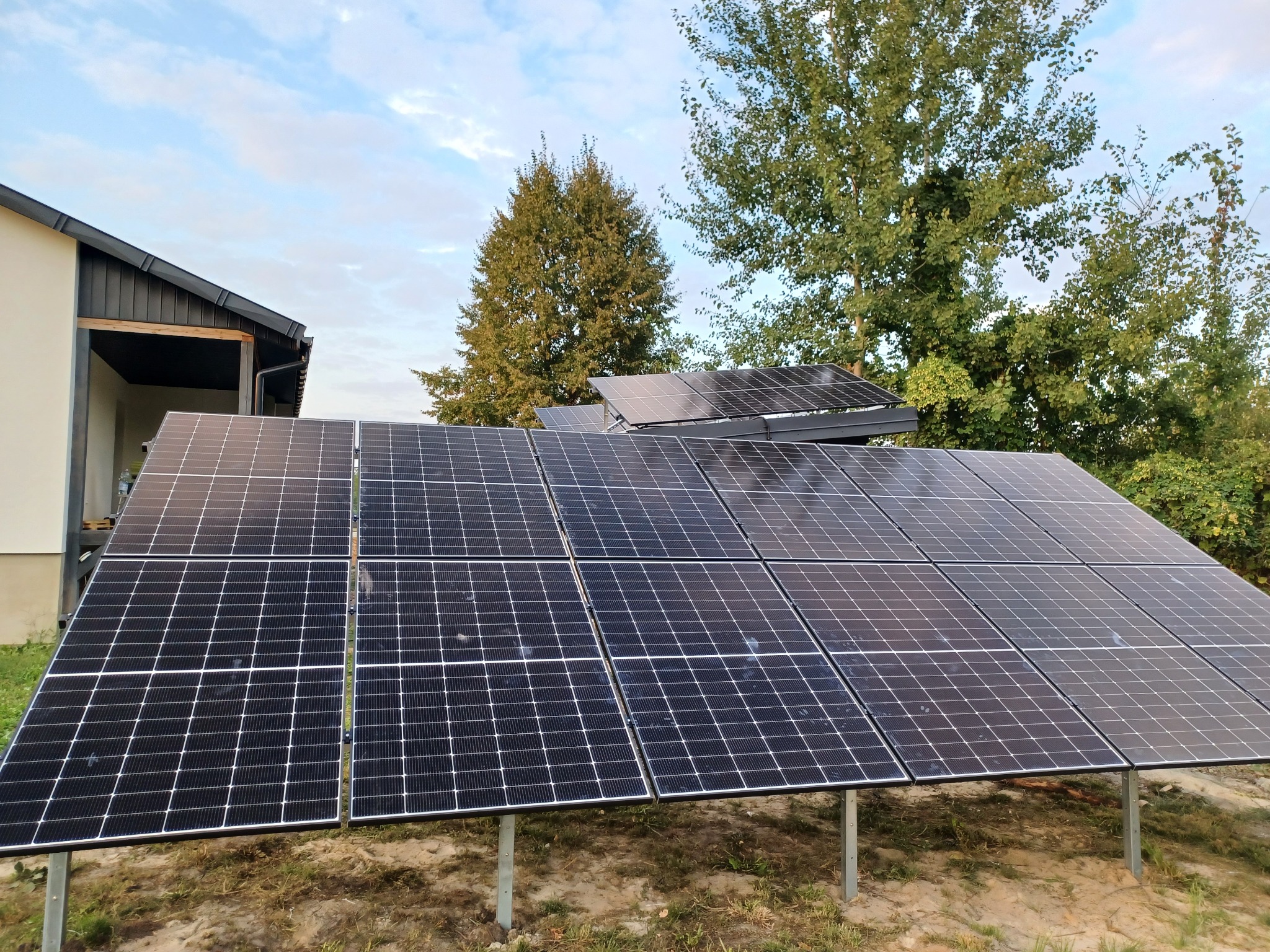 Panel słoneczny na metalowej konstrukcji przed domem jednorodzinnym, z drzewami w tle pod błękitnym niebem.