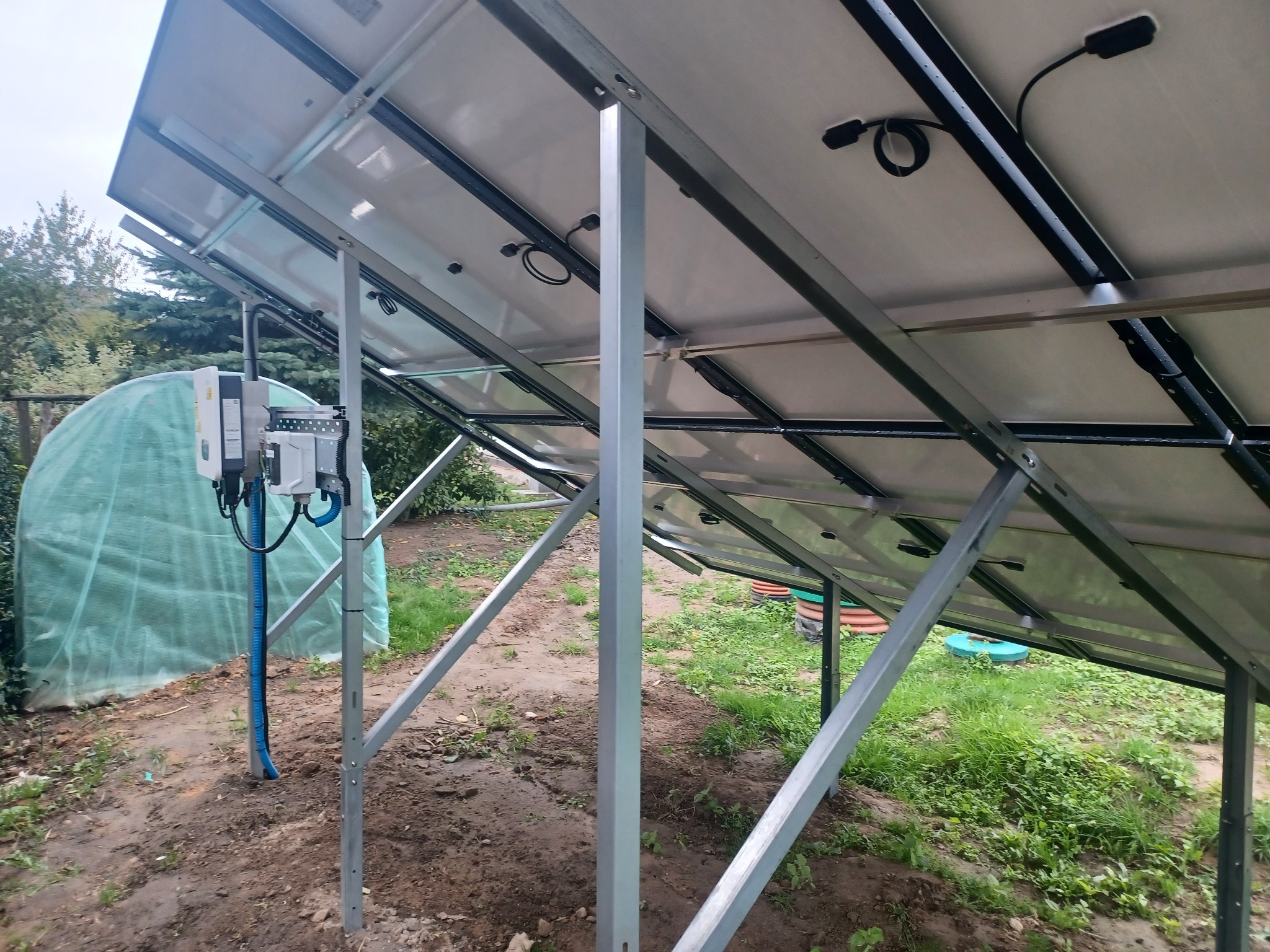 Widok od spodu na panele słoneczne zamontowane na metalowej konstrukcji, z widocznym inwerterem i okablowaniem, na tle ogrodu z tunelem foliowym.