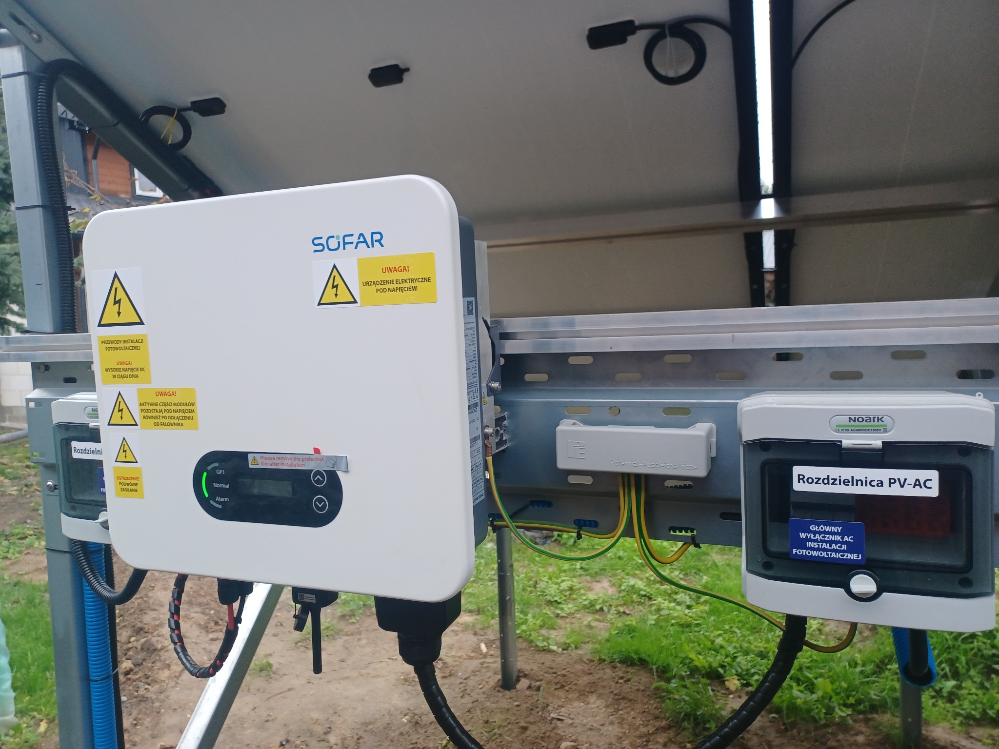 Falownik Sofar i rozdzielnica PV-AC Noark zamontowane na konstrukcji fotowoltaicznej, widoczne kable i przewody elektryczne, ostrzeżenia o wysokim napięciu.