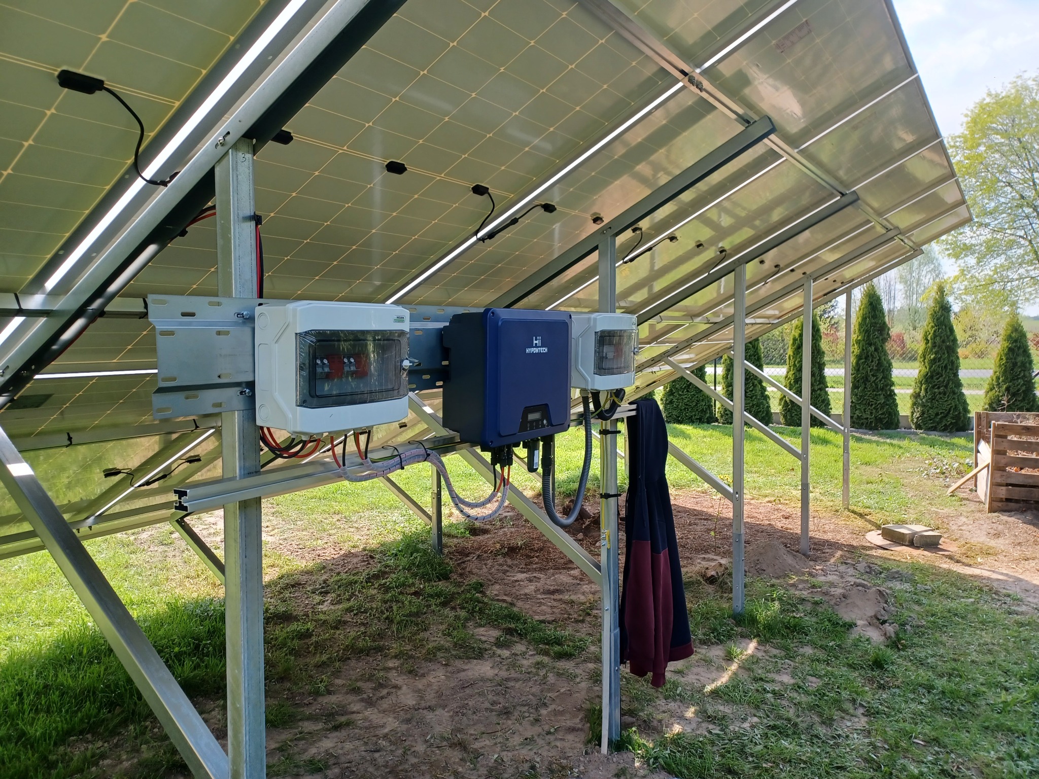 Widok od dołu na panele fotowoltaiczne zamontowane na metalowej konstrukcji, z widocznymi puszkami przyłączeniowymi i inwerterem Hypontech, w tle trawnik i tuje, na jednym ze słupków konstrukcji...