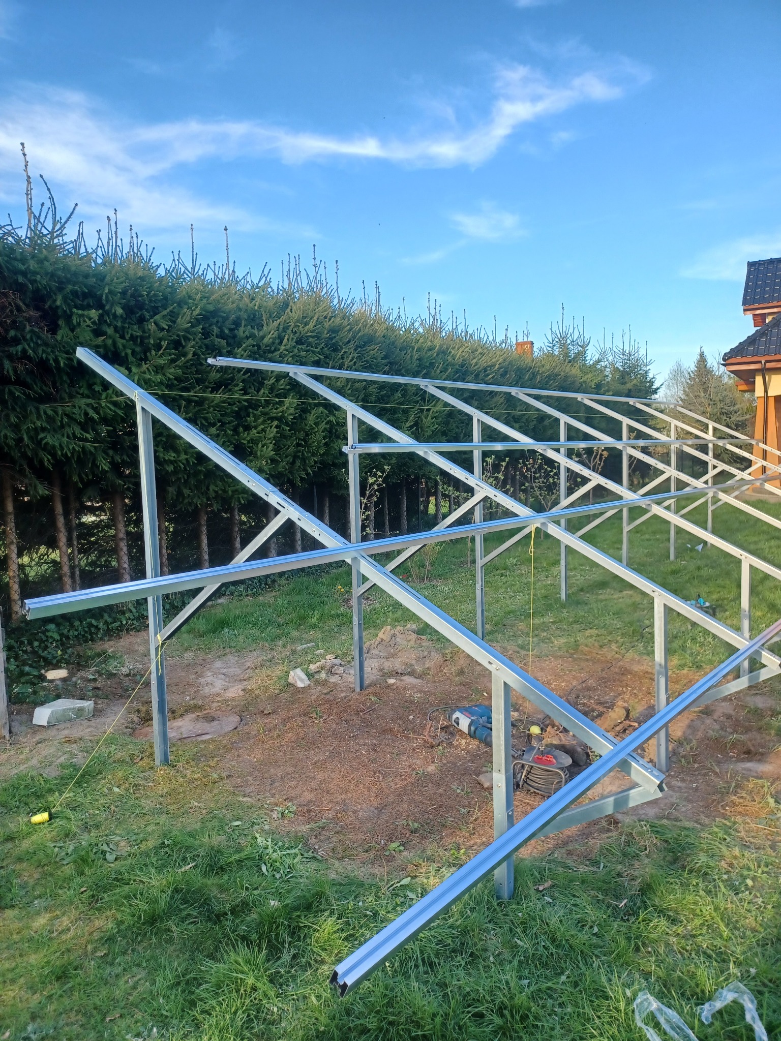 Stelaż pod panele słoneczne w trakcie montażu na trawniku przed żywopłotem, widoczne narzędzia i elementy konstrukcyjne.