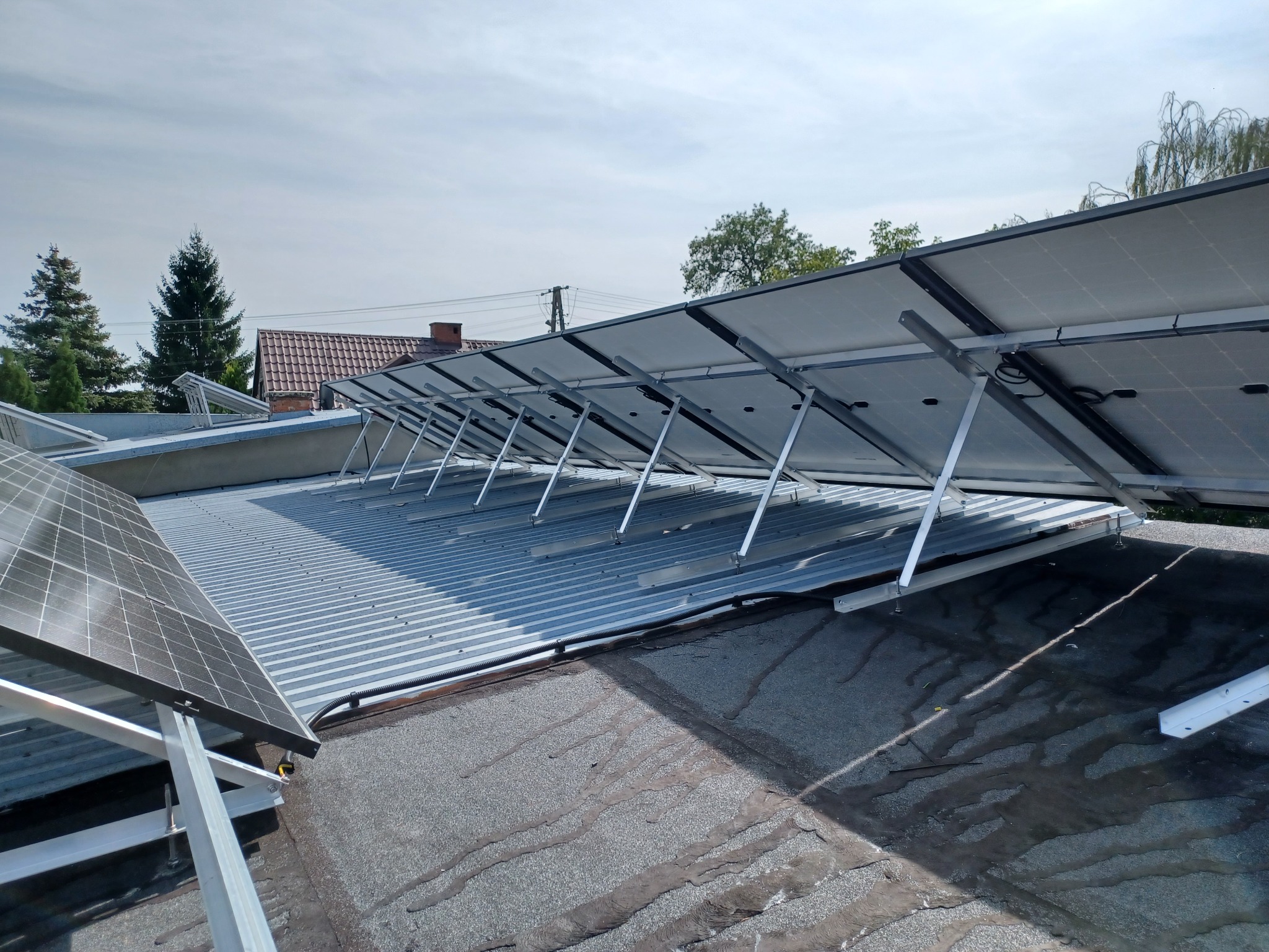 Panele fotowoltaiczne zamontowane na metalowym dachu, widoczne od strony tylnej, z widocznymi elementami montażowymi i fragmentem dachu sąsiedniego budynku w tle.