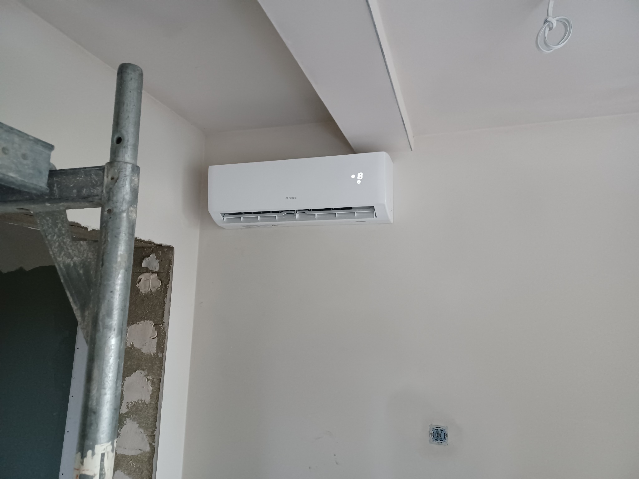 Biały klimatyzator marki Gree zamontowany na ścianie w trakcie remontu, widoczne rusztowanie i surowy tynk.