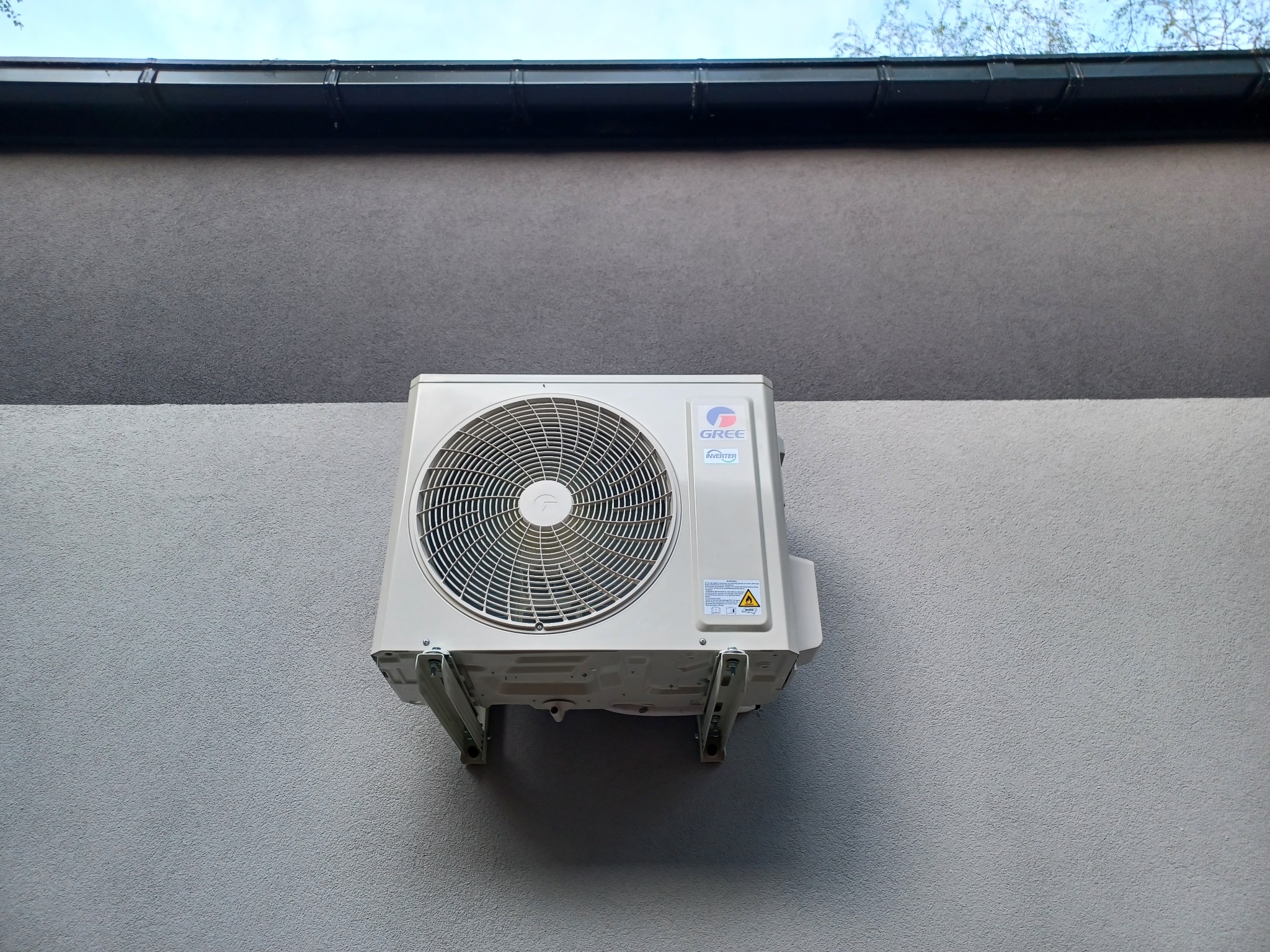 Biała jednostka zewnętrzna klimatyzatora marki Gree zamontowana na jasnoszarej elewacji dwukondygnacyjnego budynku z czarną rynną.