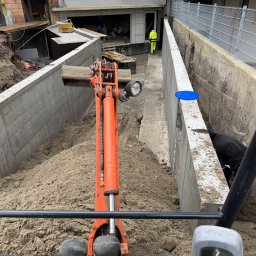 DM BUDOWNICTWO S.C. - Mini koparka w wykopie fundamentowym z betonowymi ścianami, widok z kabiny operatora. W tle pracownik w odblaskowej kamizelce.