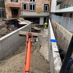 DM BUDOWNICTWO S.C. - Wykop pod fundamenty budynku wielorodzinnego z użyciem pomarańczowej koparki. Widoczny pracownik w żółtej kamizelce i fragment budynku w stanie surowym.