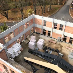 Wysoki kąt widzenia na budowę domu z czerwonej cegły i betonu, widoczne materiały budowlane, jesienne drzewa w tle.