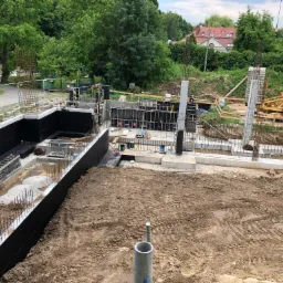 Budowa fundamentów: Wykop z izolacją pionową, szalunki i zbrojenie słupów, widoczny żuraw budowlany w tle.