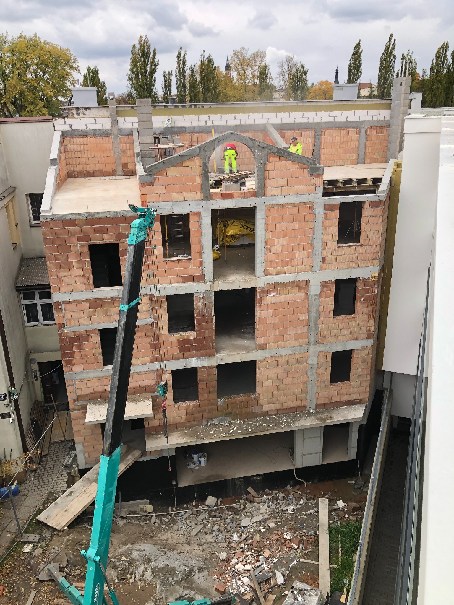 Wysoki kąt na budowę z cegły z dźwigiem. Murarze w żółtych kamizelkach pracują na górze. Widoczne okna i elementy konstrukcyjne. Pochmurne niebo w tle.