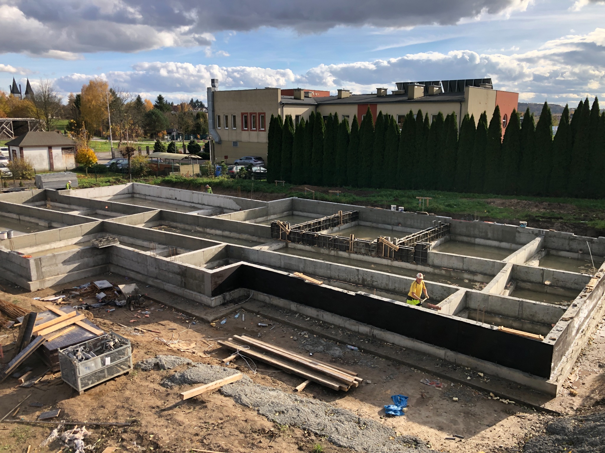 Wylane fundamenty pod budynek z widoczną hydroizolacją pionową, szalunkami i pracownikiem w żółtej koszulce. W tle budynek i drzewa iglaste.