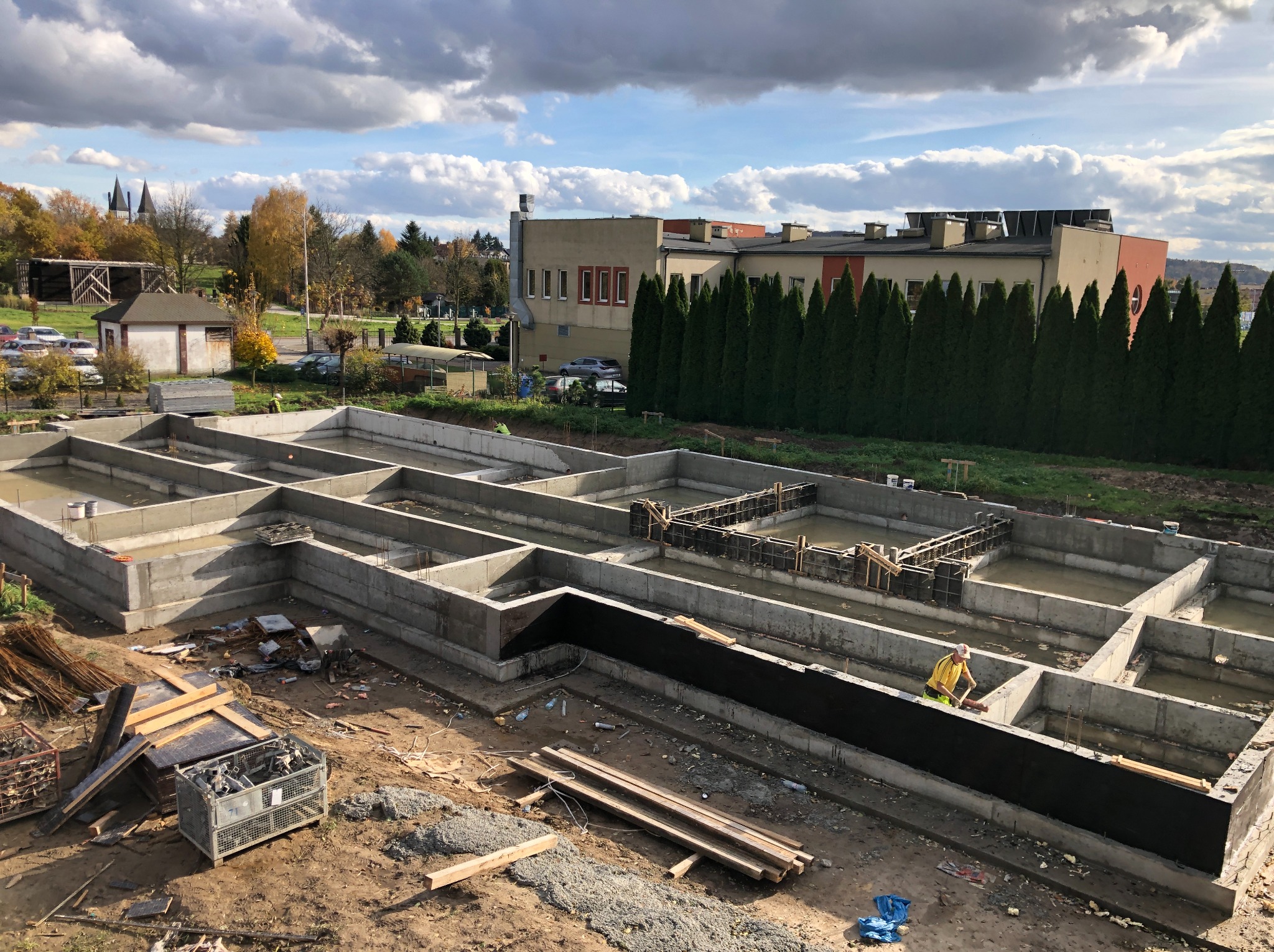 Wylane fundamenty z hydroizolacją, widok z góry na plac budowy z pracownikiem w żółtej koszulce. W tle budynki i drzewa pod pochmurnym niebem.