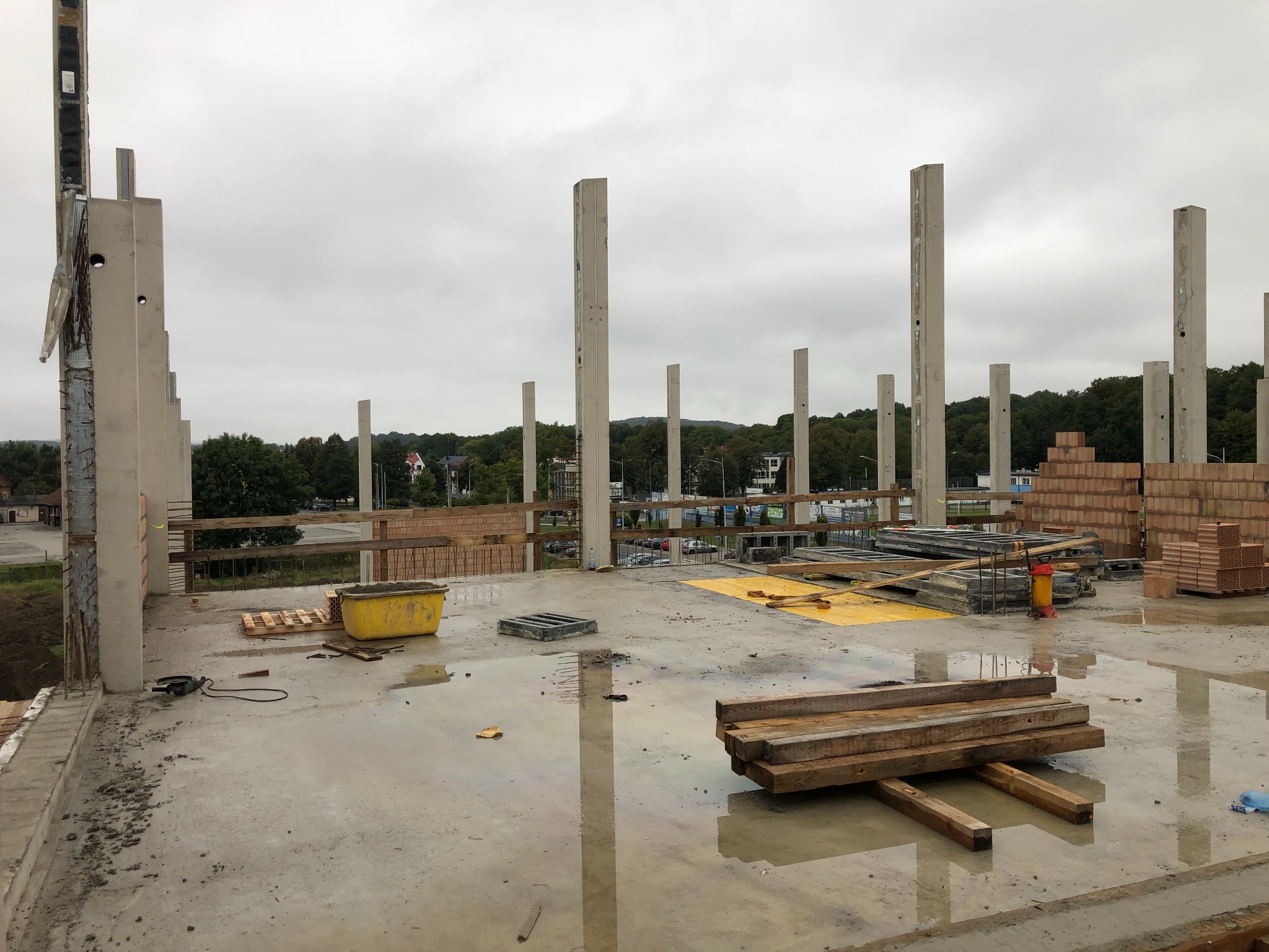 Wnętrze w trakcie budowy budynku żelbetowego z widocznymi betonowymi słupami, deskowaniem, cegłami i narzędziami. Mokra posadzka odbija elementy konstrukcji.