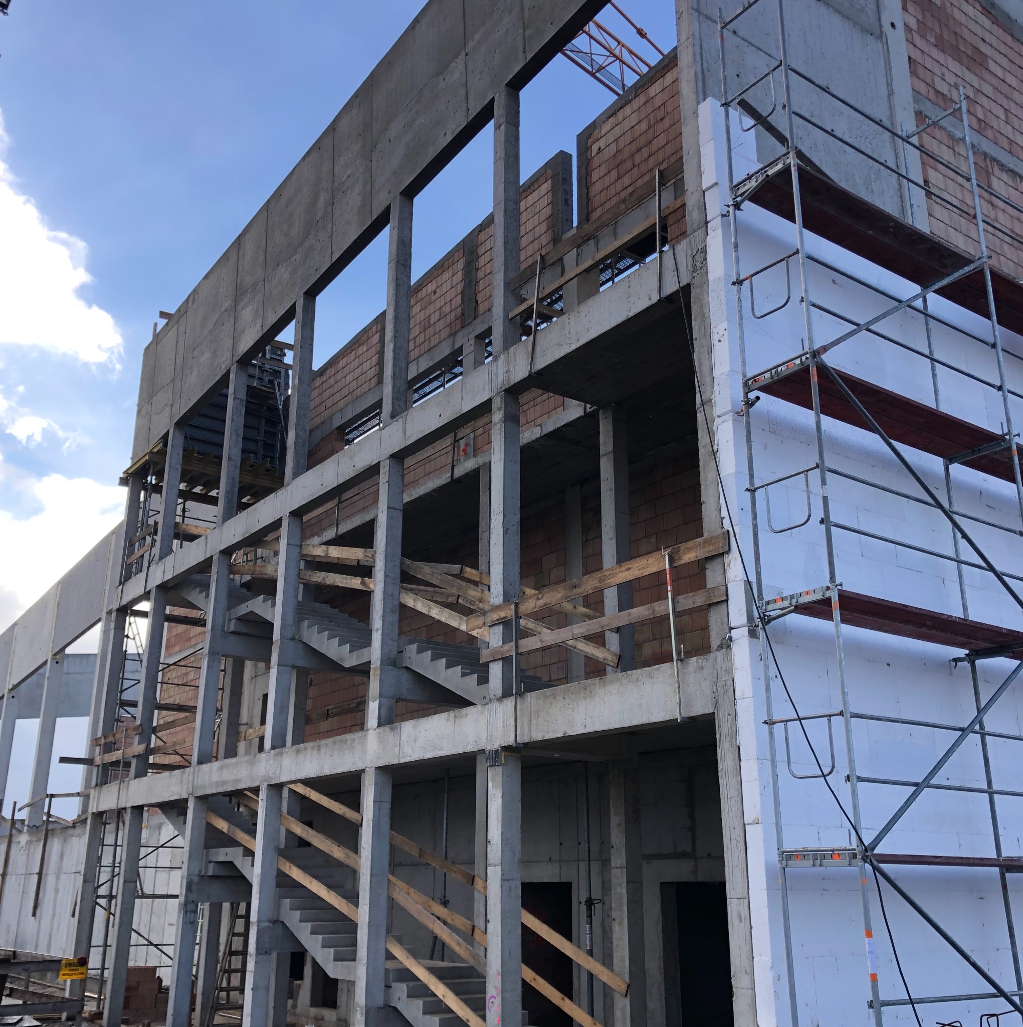 Wznoszony budynek żelbetowy z widocznymi schodami i rusztowaniami. Częściowo ocieplona ściana. Widoczne fragmenty murowanych ścian. Niebo z chmurami w tle.